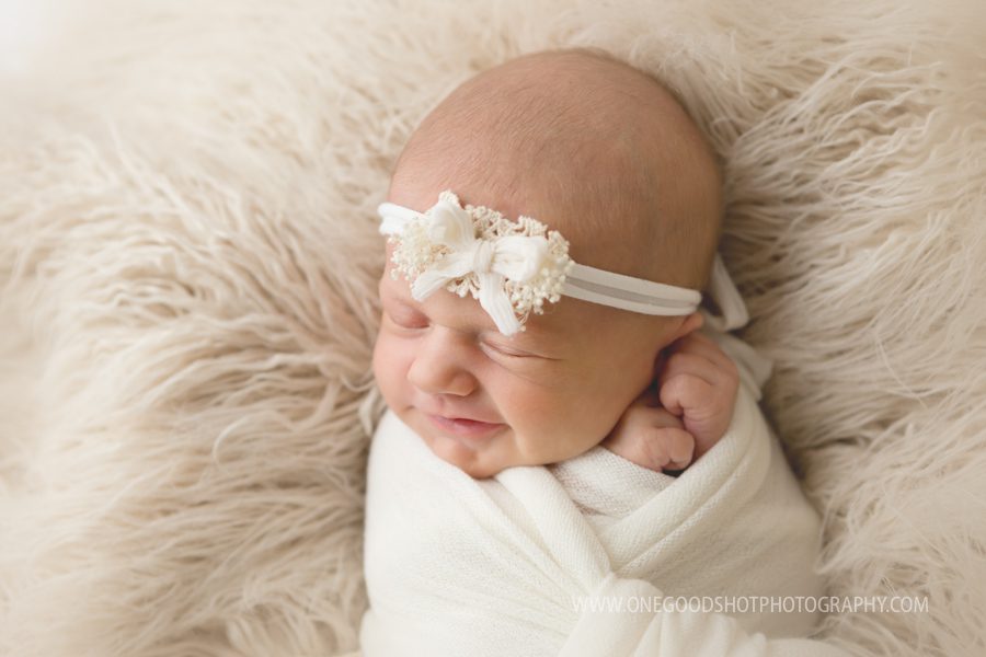 newborn baby girl wrapped in white blanket smiling with white bow headband