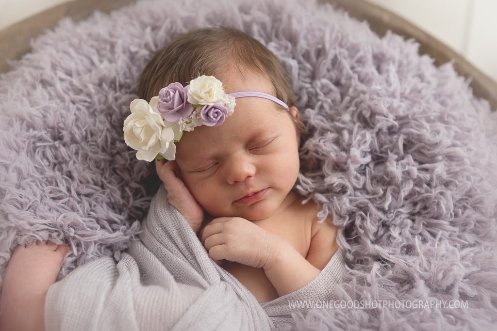 newborn girl, hand on face, wrapped in lavender on lavender fluff bowl