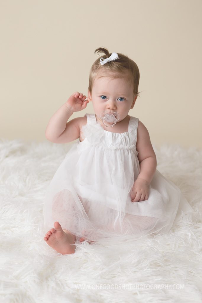 one year old baby girl sitting in a white dress with pacifier
