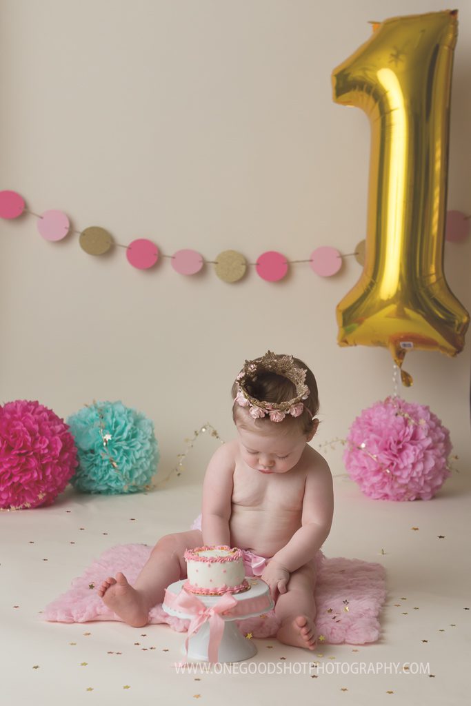 cake smash session, pink, teal, gold, one year old girl wearing princess crown, hands in cake