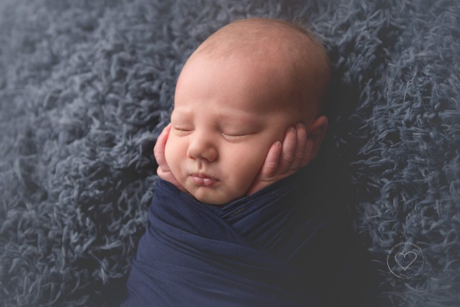 newborn boy, wrapped in navy blanket, hands on face