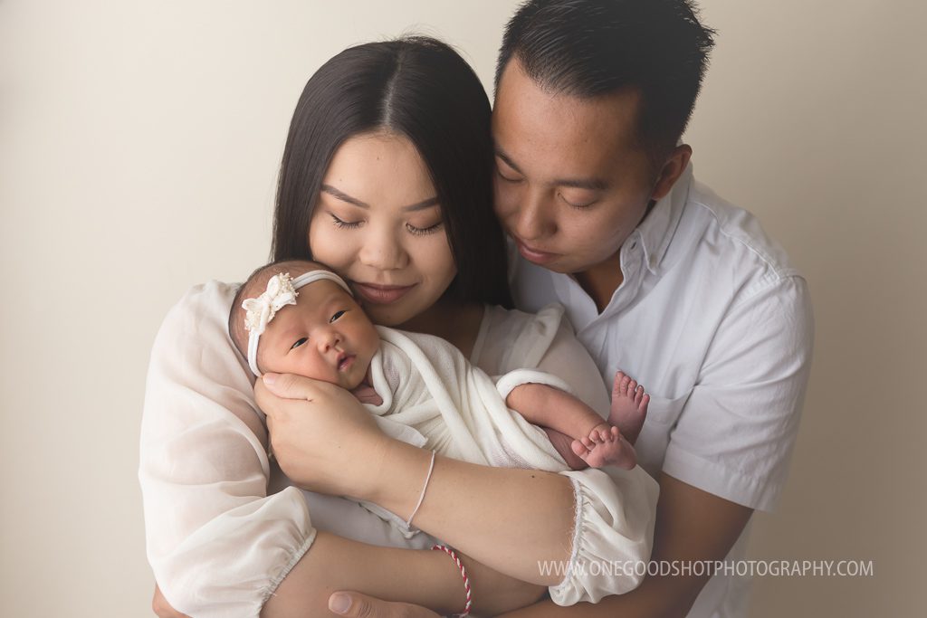newborn girl in mommy's arms with daddy wrapping around.  Cream clothing and a bone backdrop fresno photographer