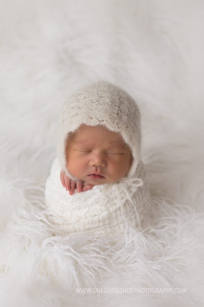 newborn girl wearing white bonnet in a potato sack pose on white fur fresno photographer