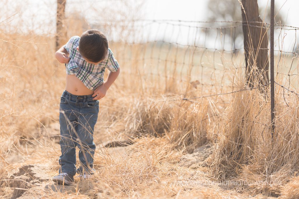 boy in field, looking at belly button, cutting hands in pockets, rustic fence