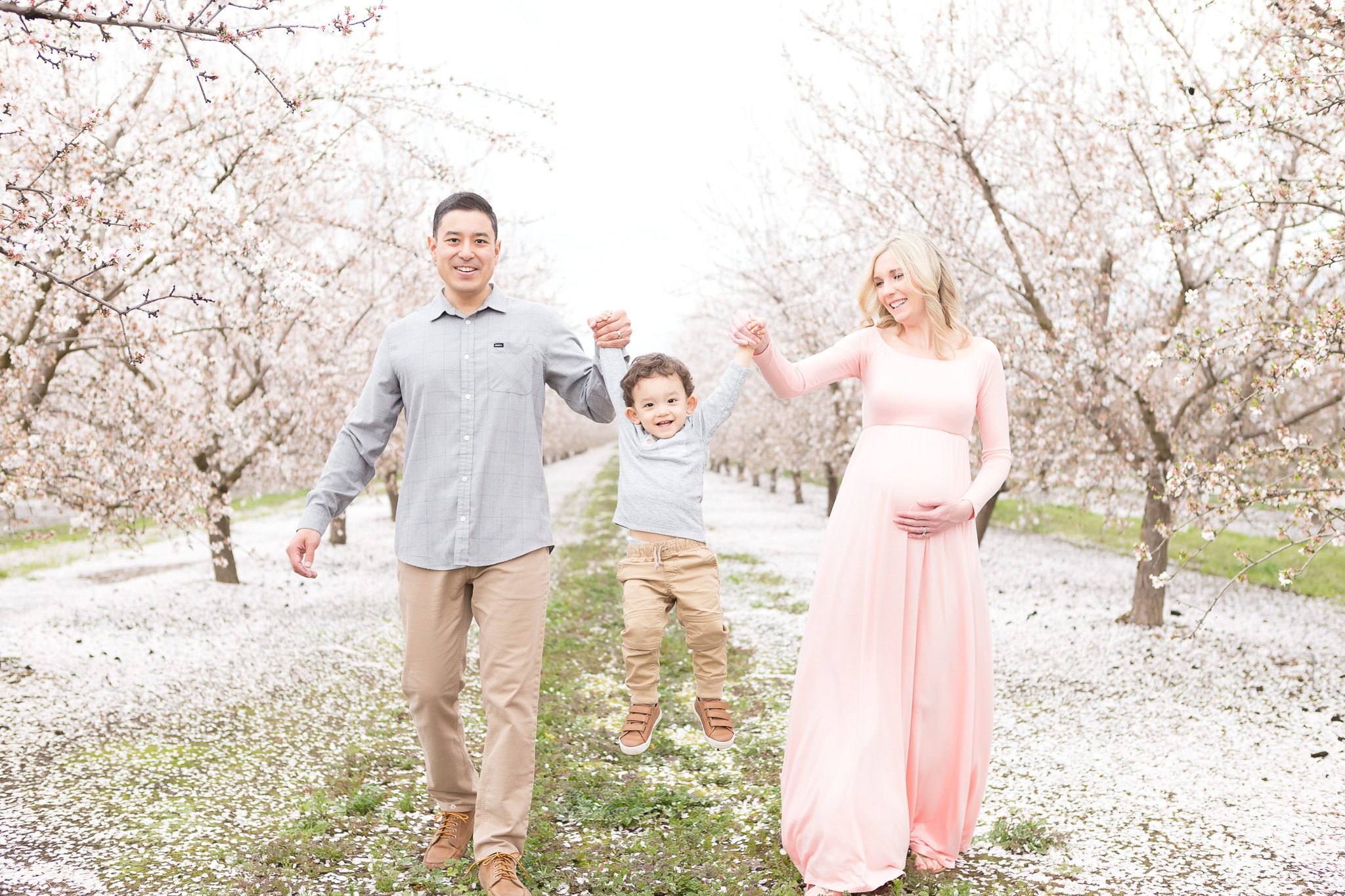 maternity session, pregnancy photos, pink maternity dress, mom and dad walking in the blossoms swinging toddler