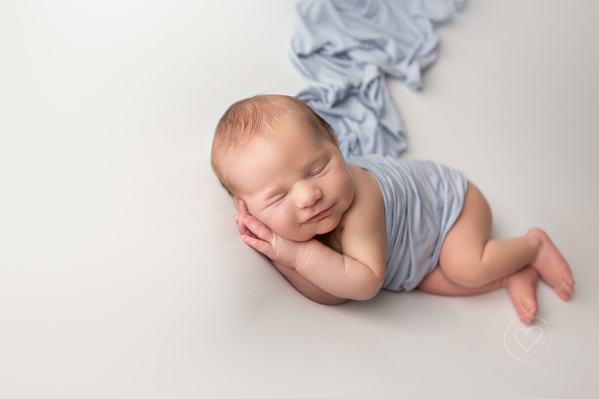 Newborn boy, side lying, white backdrop, blue wrap, smiling
