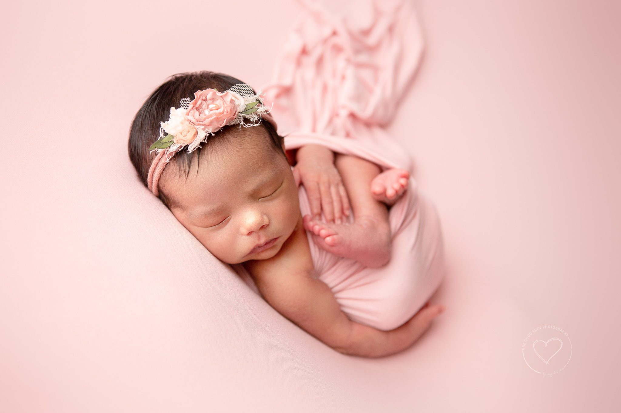 Newborn girl, huck Finn pose, wrapped in pink on pink backdrop, wearing pink floral headband