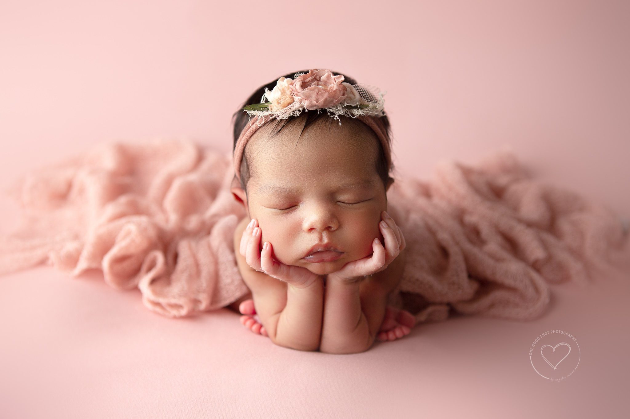 Newborn girl in froggy pose on pink backdrop, wearing pink floral headband
