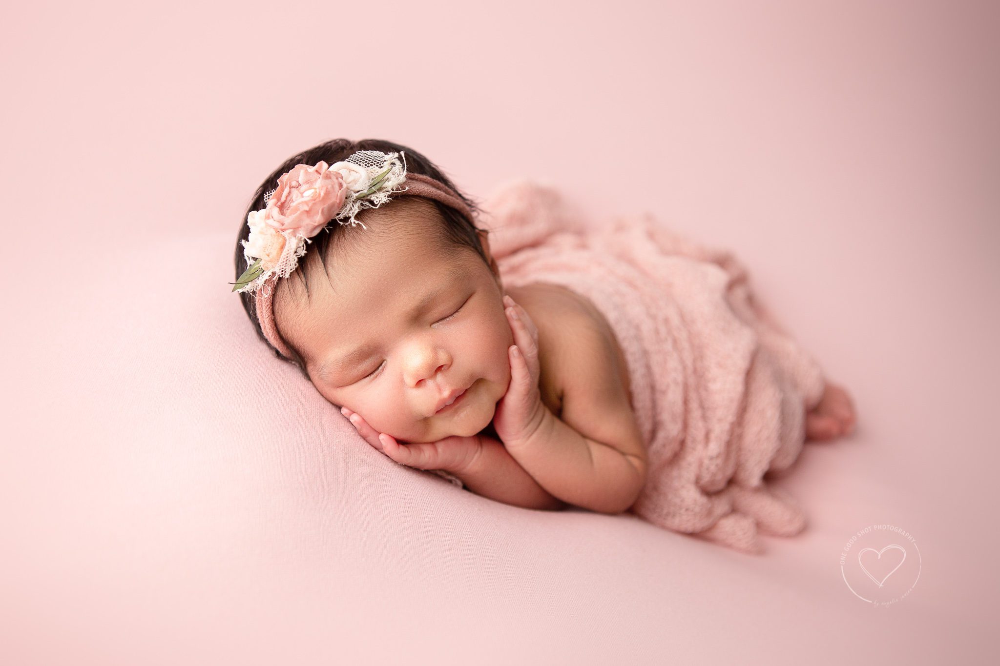Newborn girl in side lying pose with hands on cheeks.  Pink wrap, pink backdrop, pink floral headband