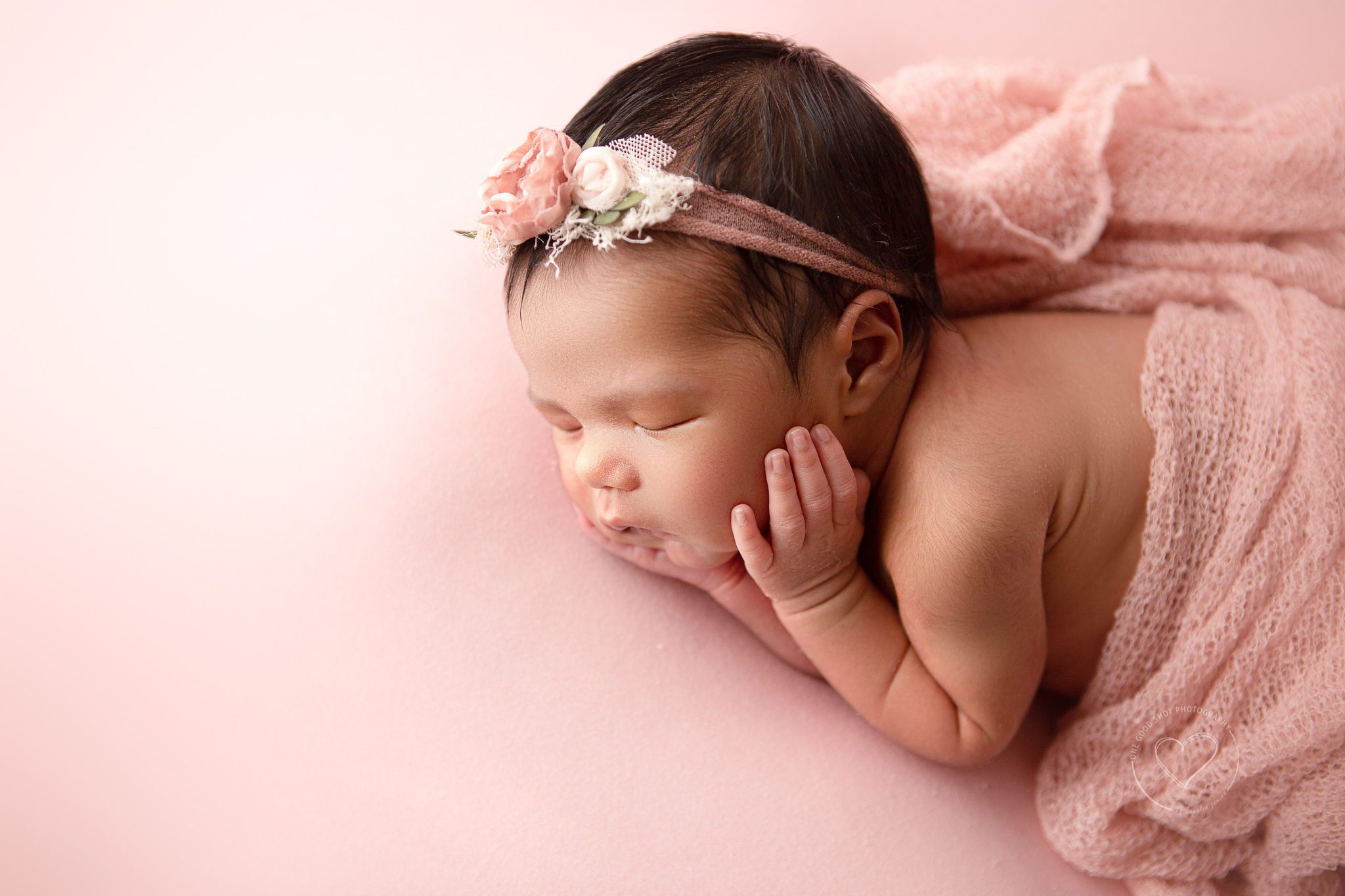 Newborn girl in side lying pose with hands on cheeks.  Pink wrap, pink backdrop, pink floral headband, profile