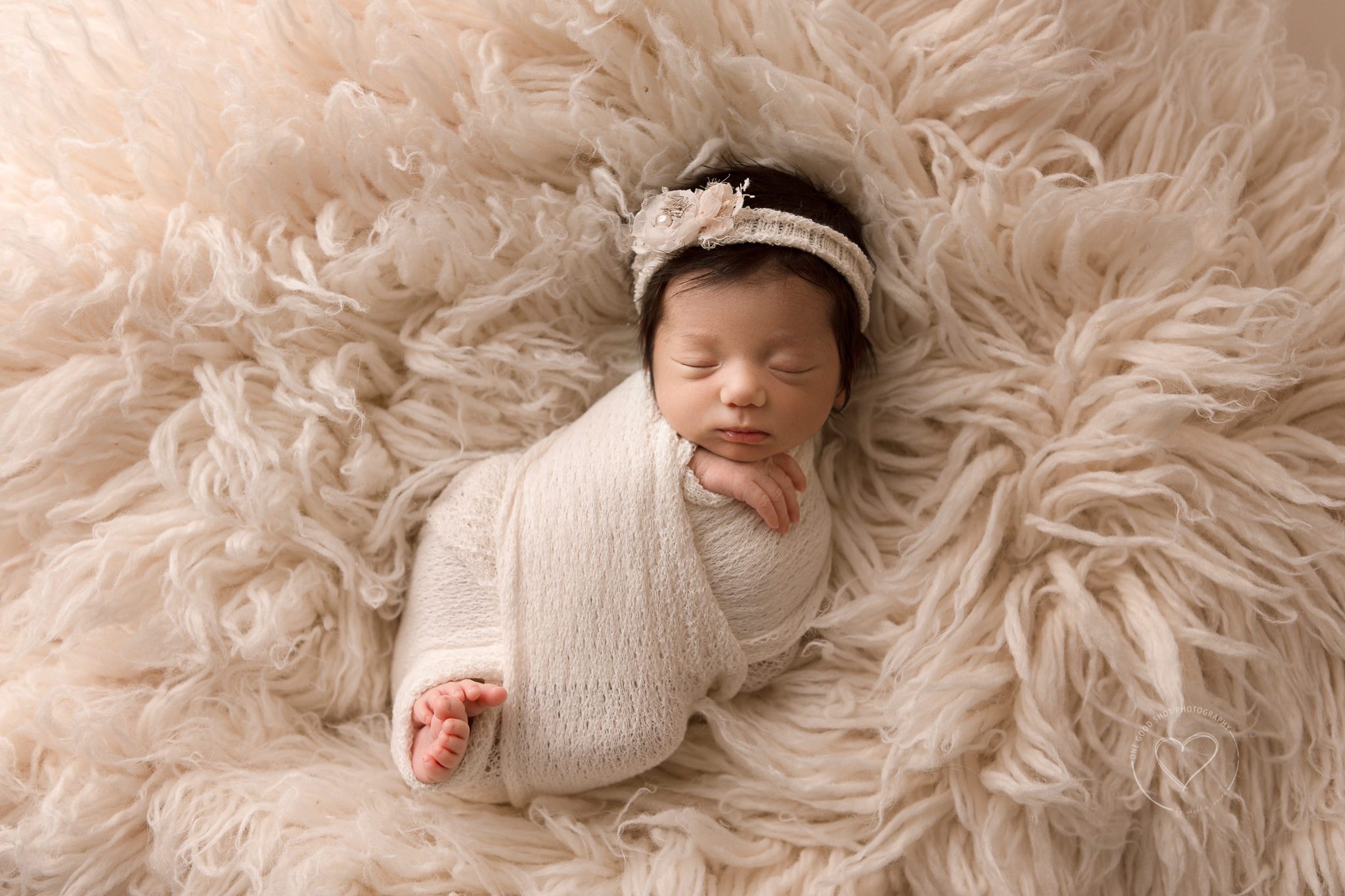 Fresno newborn baby girl photo, wrapped in cream, hands and get showing, wearing floral headband, lying on flokati rug