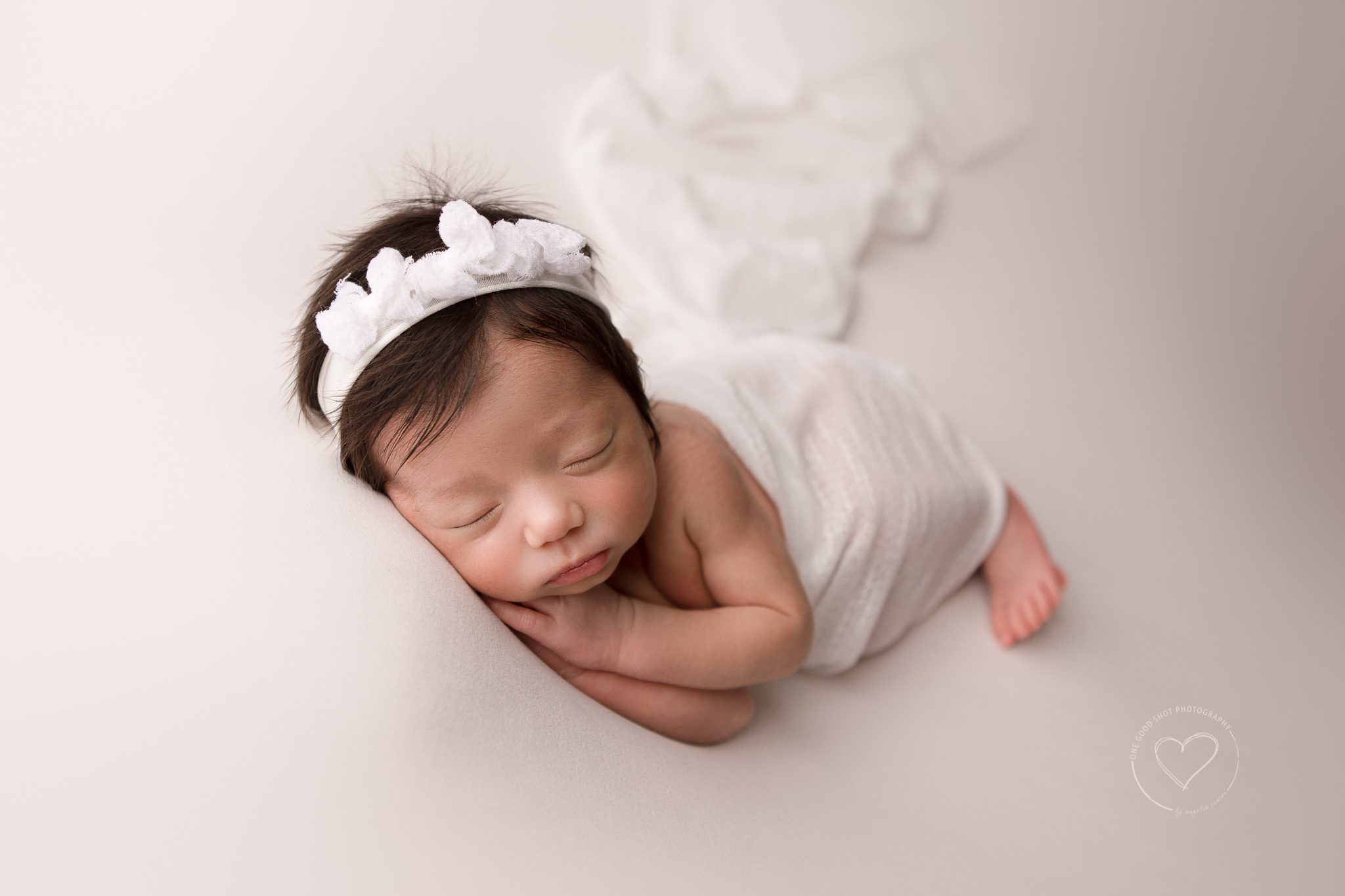 Fresno Newborn Photographer, Baby girl side lying, hands under cheek, covered with white layer on white backdrop, wearing white bow headband