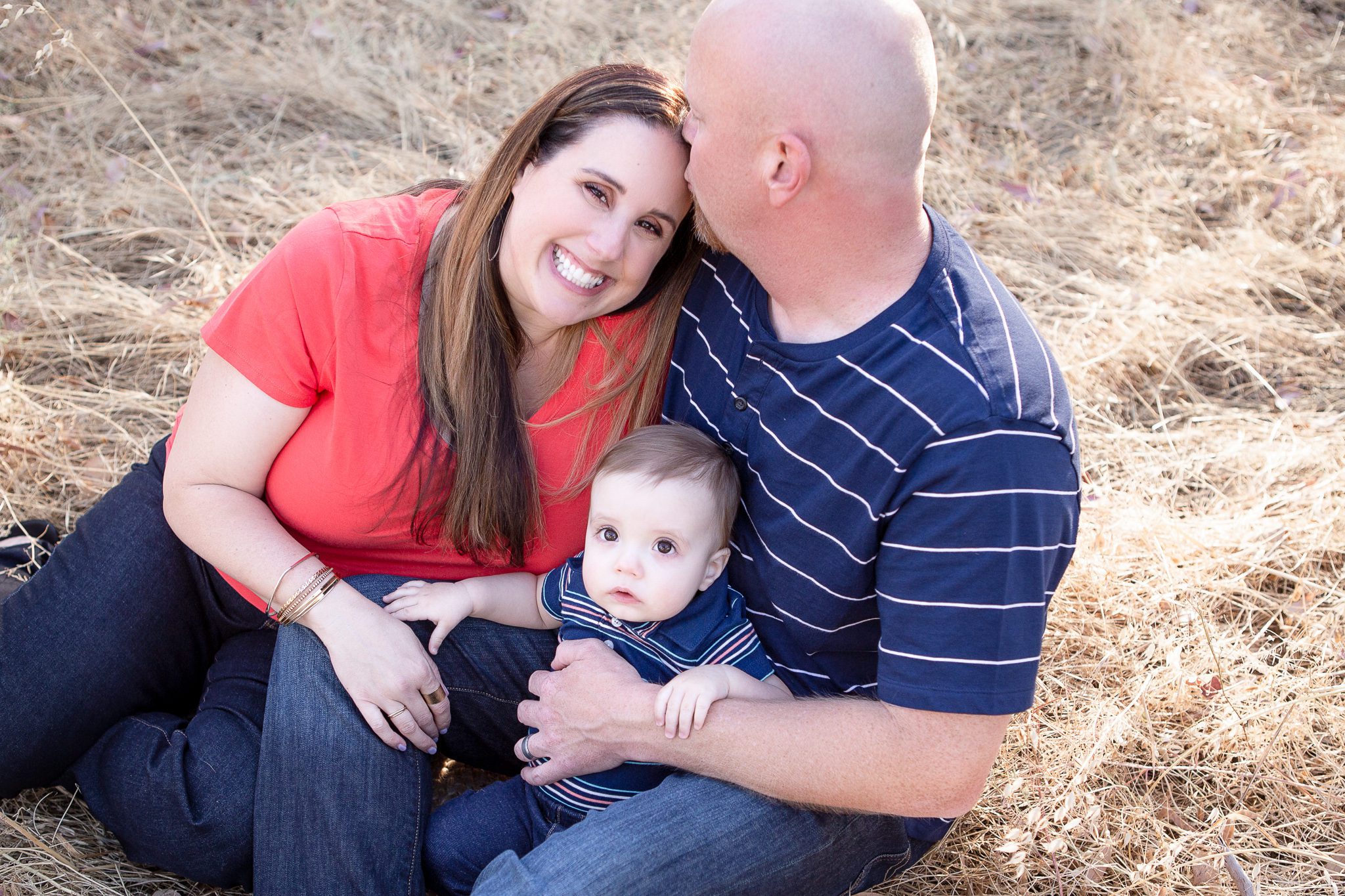 Fresno Family Photographer, sitting in a field, dad kissing mom, mom and baby smiling