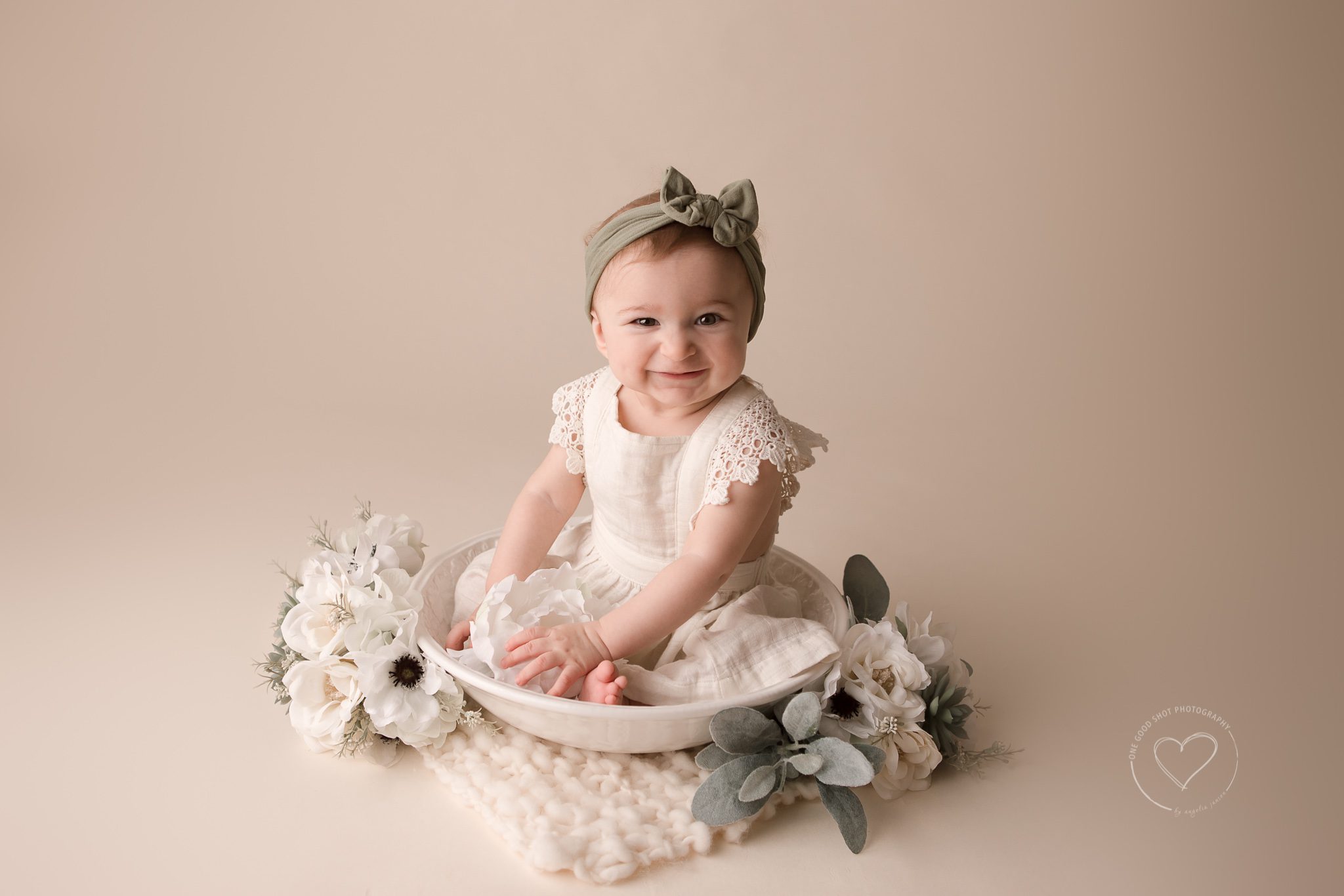 Baby Girl, 6 Months, Sitter Session, Baby wearing white linen dress and a green bow sitting in a bowl with flowers smiling at the camera