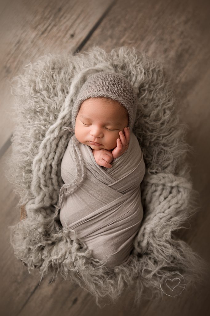 newborn boy, wrapped in gray, laying in basket with gray layers, hand on face, bonnet